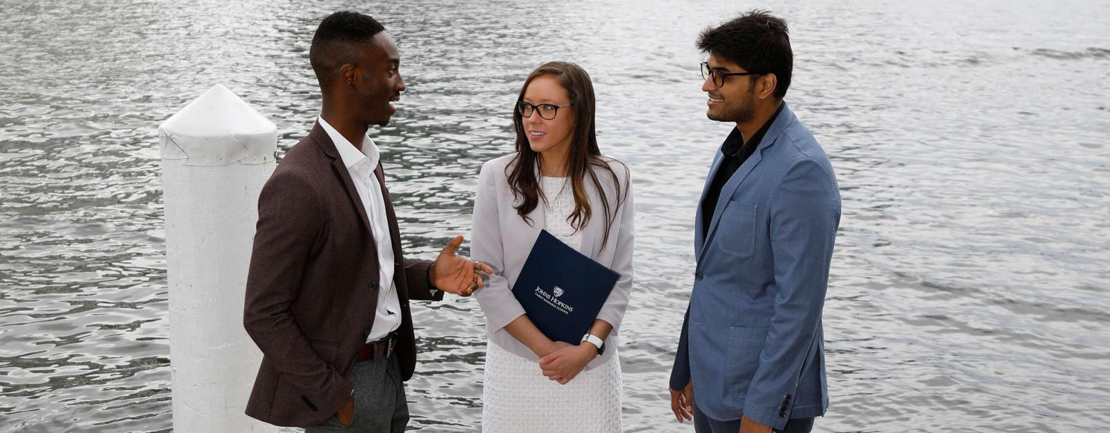 3 students near the water networking and holding a JHU Carey folder
